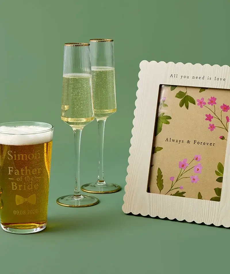 A personalised beer glass, two champagne flutes, and a decorative photo frame with floral artwork arranged on a green background. A personalised beer glass, two champagne flutes, and a decorative photo frame with floral artwork arranged on a green background.
